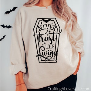 May include: A beige sweatshirt with a black graphic print of a coffin with the text "Never trust the living" and a bat inside the coffin.