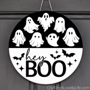 Op de afbeelding: Ronde Halloween-bord met een zwart-wit ontwerp. De bovenste helft heeft witte spookillustraties op een zwarte achtergrond. De onderste helft is wit met de woorden "hey BOO" in zwart, samen met vleermuis- en sterrenafbeeldingen.