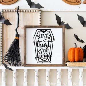 May include: A wooden sign with a black silhouette of a coffin with the text "Never trust the living" inside. The sign is on a white shelf with a black broom and a small orange pumpkin.