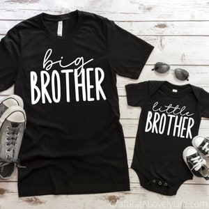 Op de afbeelding: Twee zwarte T-shirts met witte tekst. Het grotere shirt zegt "grote broer" en het kleinere shirt zegt "kleine broer".
