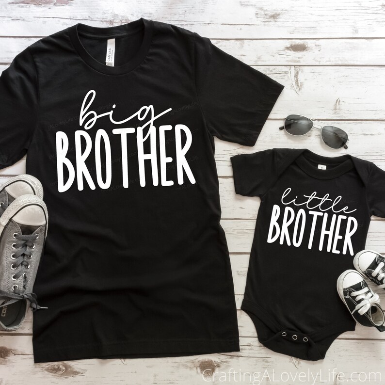 Op de afbeelding: Twee zwarte T-shirts met witte tekst. Het grotere shirt zegt "grote broer" en het kleinere shirt zegt "kleine broer".