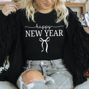 May include: Black crew neck t-shirt with the words "happy NEW YEAR" in white script and a bow graphic. The shirt is worn with a black cardigan and light wash jeans.