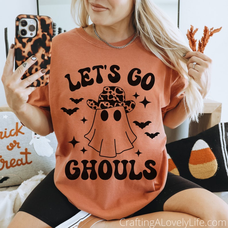 Może przedstawiać: Pomarańczowy t-shirt z czarnym nadrukiem ducha w kowbojskim kapeluszu w panterkę. Pod duchem znajduje się napis "Let's Go Ghouls" w kolorze czarnym.