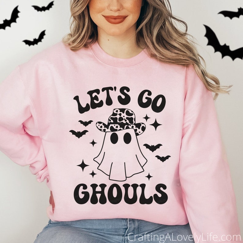 Może przedstawiać: R&oacute;żowa bluza z czarnym nadrukiem ducha w kowbojskim kapeluszu z wzorem krowy. Na bluzie nadrukowany jest czarny napis "Let's Go Ghouls".