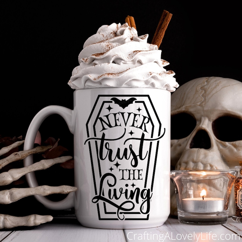 May include: White coffee mug with a black graphic of a coffin with the text "Never trust the living" and a bat inside the coffin. The mug has whipped cream on top with cinnamon sprinkled on it.