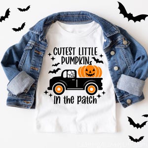 May include: White t-shirt with a black and orange pumpkin design. The pumpkin is in a black truck with a spiderweb on the side. The text on the shirt reads "Cutest Little Pumpkin in the Patch".