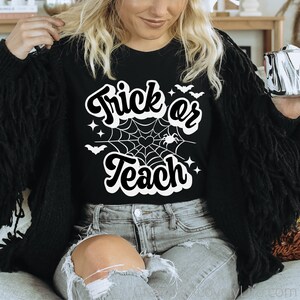 Peut inclure: Un t-shirt noir avec un motif blanc qui dit "Trick or Teach" avec une toile d'araign&eacute;e et des chauves-souris.