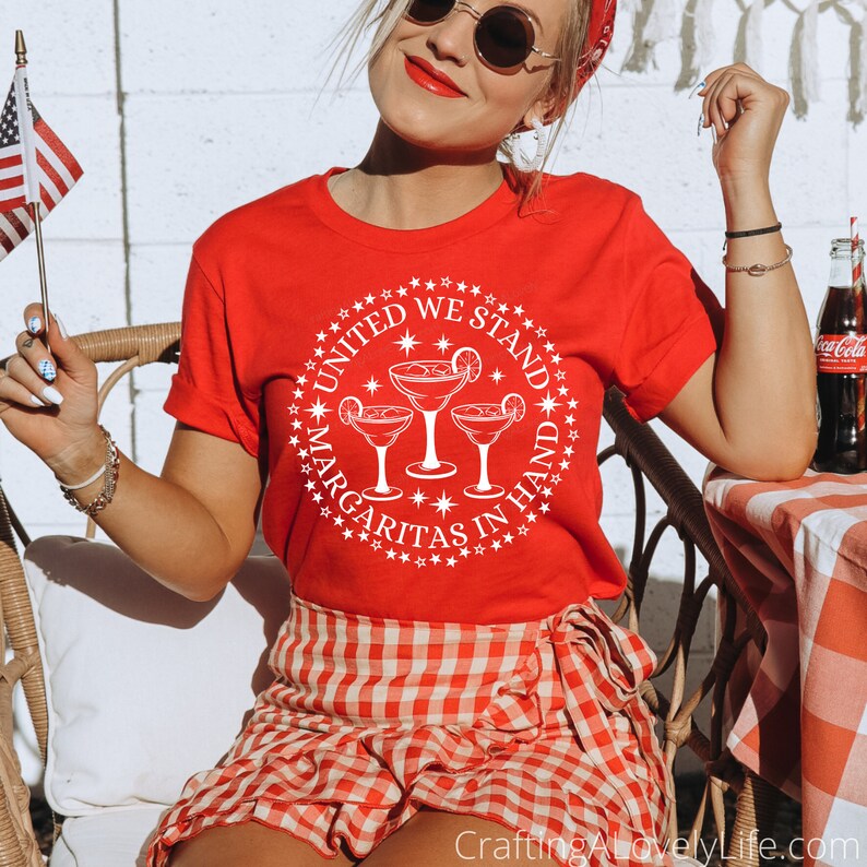 May include: A red t-shirt with a white circle design featuring the text "United We Stand Margaritas in Hand" and three margarita glasses. The design also includes stars and small white circles.