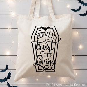 May include: A white canvas tote bag with a black graphic of a coffin with a bat and the text "Never trust the living".