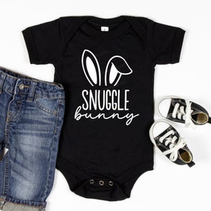 Puede incluir: Mono negro para beb&eacute; con orejas de conejo blancas y el texto "Snuggle Bunny". El mono se combina con pantalones cortos de mezclilla azul y zapatillas negras.