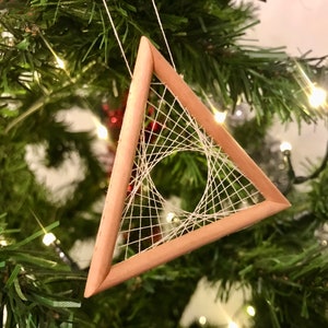 May include: A wooden triangle Christmas ornament with a white string design. The ornament is hanging on a green Christmas tree with small white lights. The ornament is made of light brown wood.