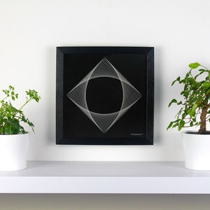 May include: A black framed string art piece with a geometric design, featuring a square shape with a central circular void. Two potted green plants flank the artwork on a white shelf, creating a minimalist aesthetic.