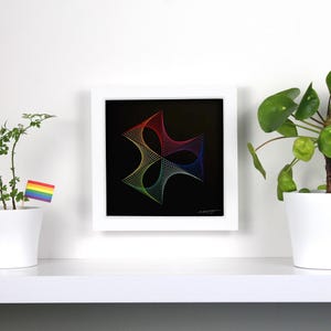 May include: A framed artwork with a geometric design in rainbow colors hangs on a white wall. Two potted plants flank the artwork, one with a small rainbow flag. The artwork features a black background.