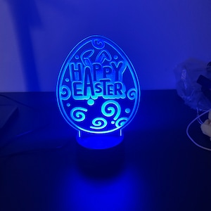 Peut inclure: Une lumière bleue éclaire une pancarte en acrylique blanche en forme d'œuf avec les mots "Joyeuses Pâques" et des volutes décoratives.