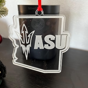 Può includere: Un ornamento natalizio in acrilico trasparente a forma dello stato dell'Arizona con le lettere "ASU" e una punta di lancia stilizzata all'interno. L'ornamento è dotato di un nastro rosso.