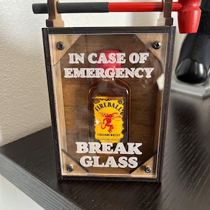 May include: A wooden box with a glass front containing a small bottle of Fireball Cinnamon Whisky. The box has a red hammer attached to the top and the text "IN CASE OF EMERGENCY BREAK GLASS" printed on the front.