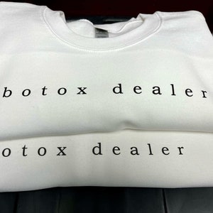 May include: Two white crewneck sweatshirts stacked on top of each other. The text "botox dealer" is printed in black on the front of both sweatshirts.