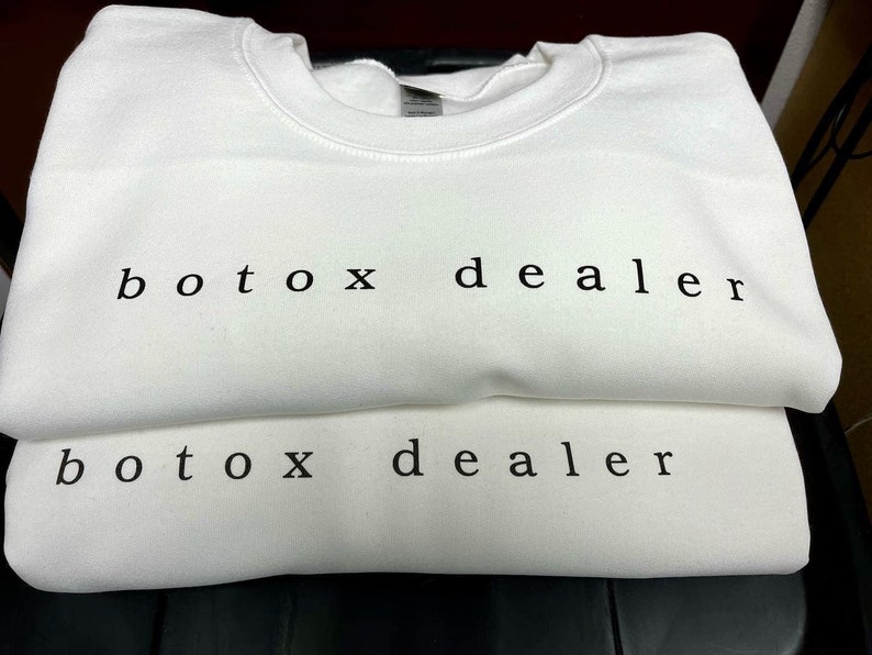 May include: Two white crewneck sweatshirts stacked on top of each other. The text "botox dealer" is printed in black on the front of both sweatshirts.