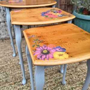 May include: A set of three nesting tables. The tables have light blue painted legs and wooden tops. The tops are decorated with floral designs in various colors. The tables are arranged in a nested configuration.