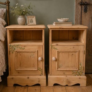 May include: A pair of wooden bedside cabinets in a natural wood finish. Each cabinet features a single door with a floral motif and a round knob. The cabinets are positioned on a wooden floor. One has a vase of flowers and a framed picture on top.