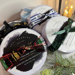 May include: Several round candles with decorative ribbons. One candle has a black and gold ribbon, another has a Dallas Cowboys ribbon, and a third has a green velvet ribbon. The labels read "JOZI CANDLES" and list essential oils and net weight 16 oz.