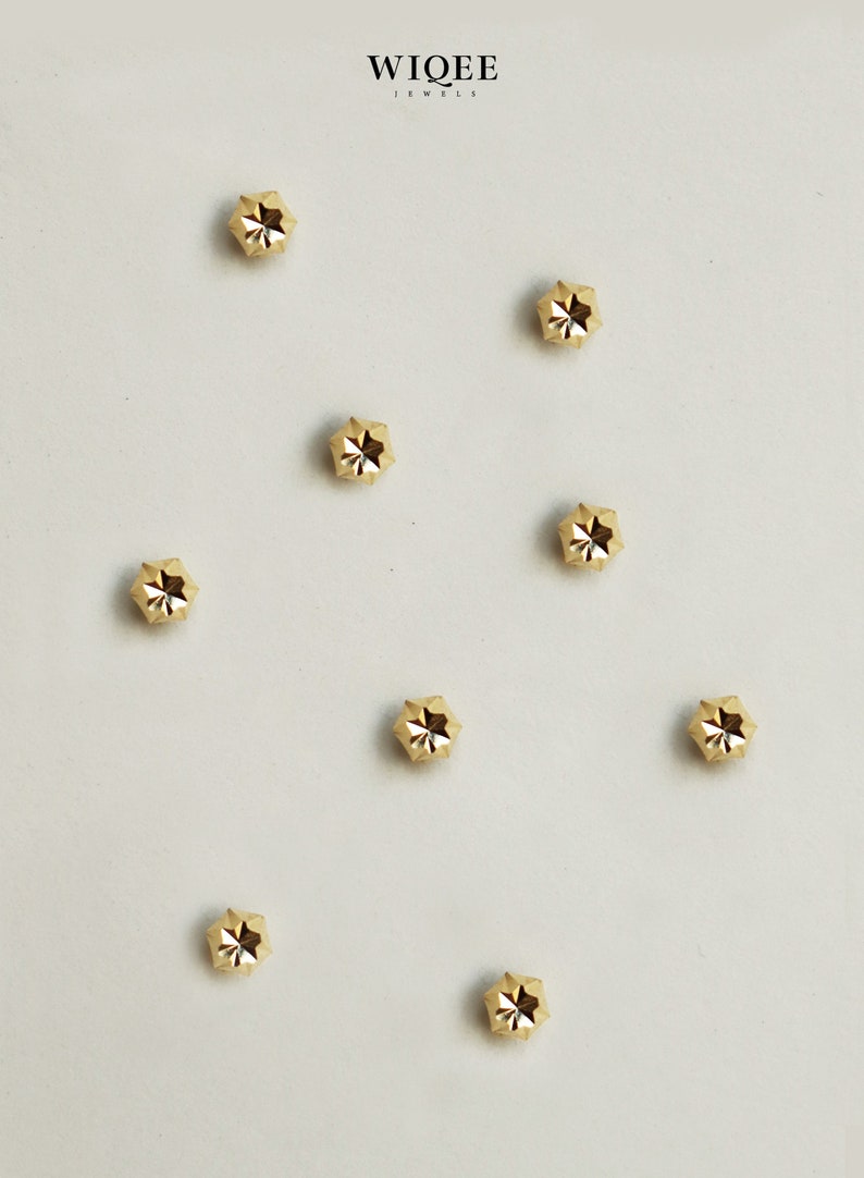Puede incluir: Ocho tachuelas doradas en forma de estrella sobre un fondo blanco. Las tachuelas son peque&ntilde;as y tienen un dise&ntilde;o facetado.