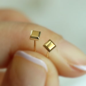 May include: A pair of gold earrings, one square and one diamond shaped. The earrings are on a person's finger.
