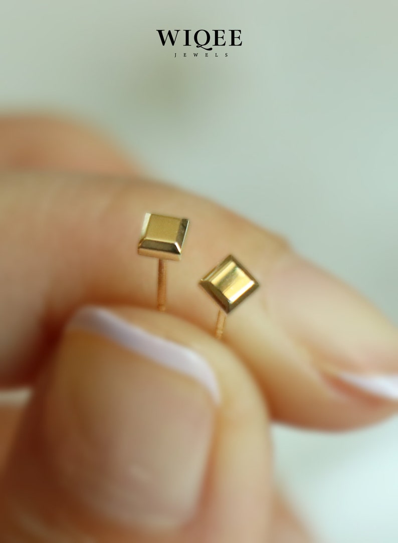 May include: A pair of gold earrings, one square and one diamond shaped. The earrings are on a person's finger.