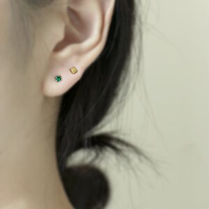 May include: A close-up of a person's ear with two small earrings. The earring on the left is a square-shaped green gemstone, and the earring on the right is a small gold square.