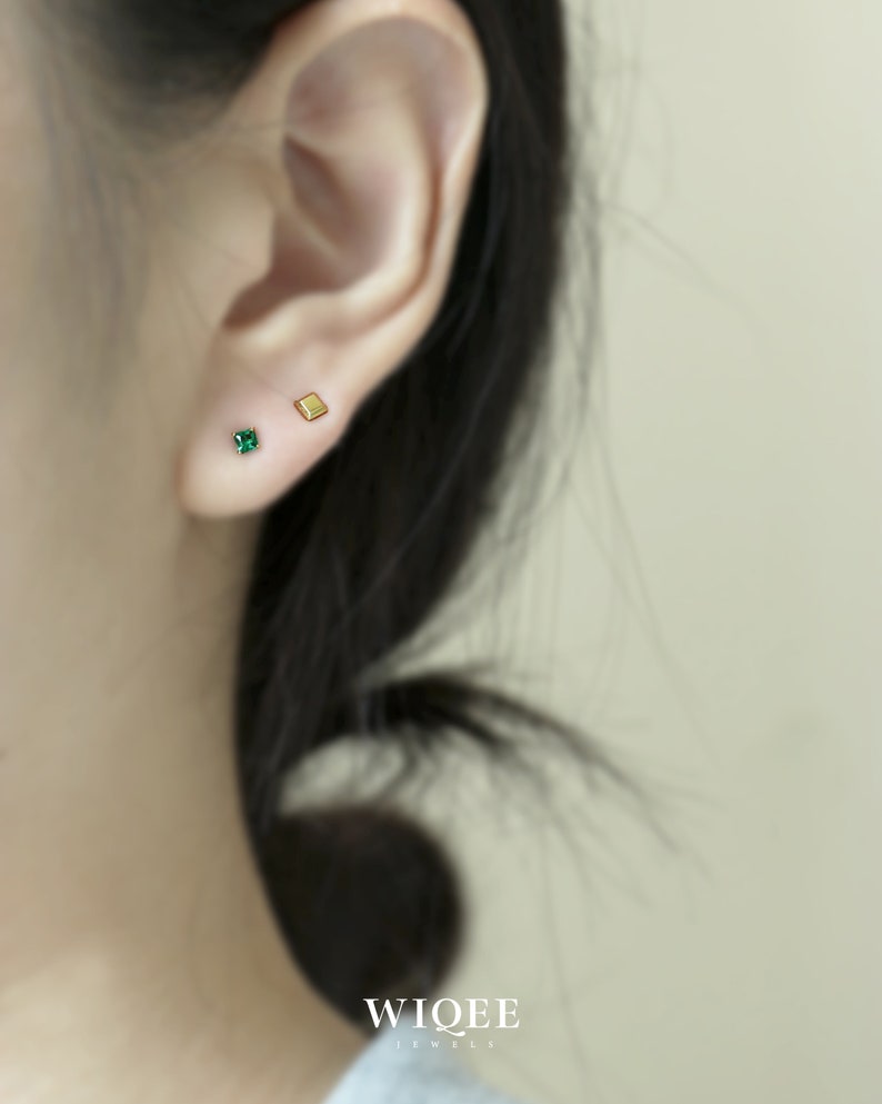 May include: A close-up of a person's ear with two small earrings. The earring on the left is a square-shaped green gemstone, and the earring on the right is a small gold square.