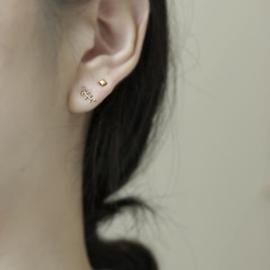May include: A close-up of a person's ear with two gold earrings. The earrings are a small square and a delicate branch design.