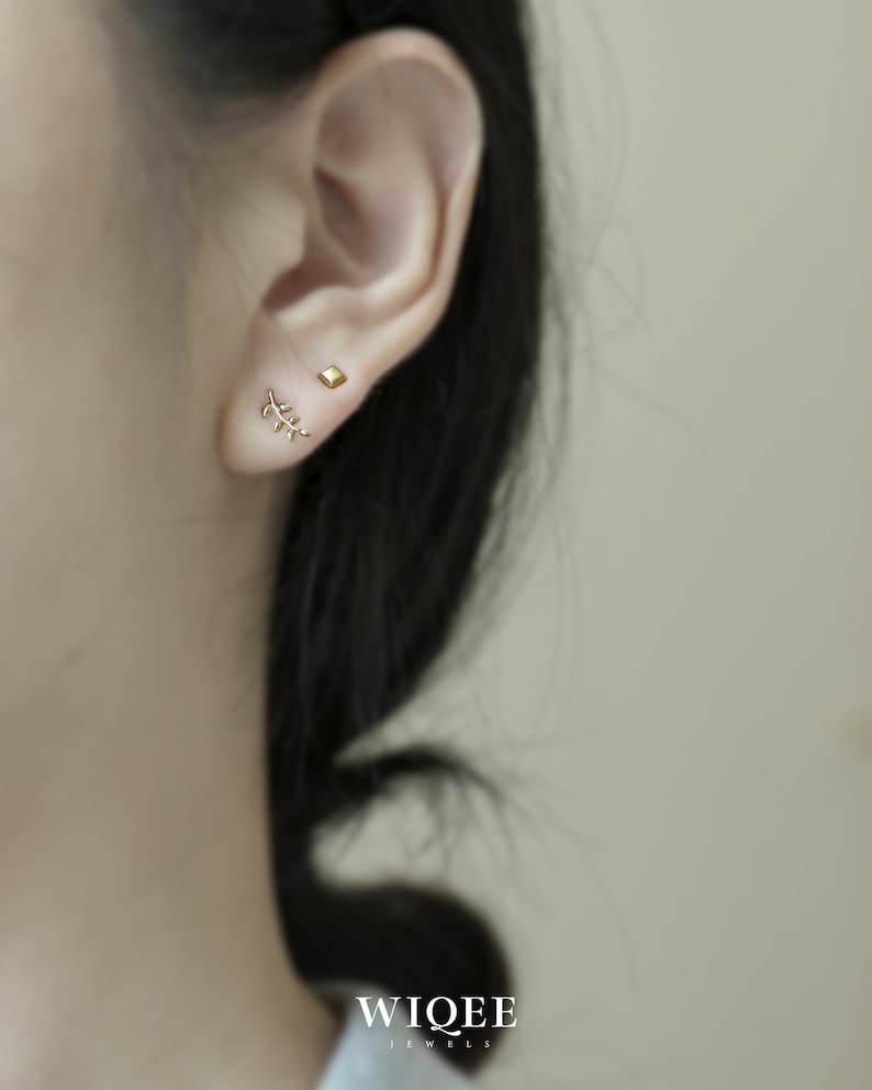 May include: A close-up of a person's ear with two gold earrings. The earrings are a small square and a delicate branch design.