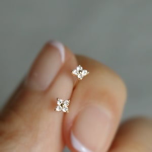 以下が含まれることがあります： 小さな花のデザインで、透明なダイヤモンドがセットされた、ゴールドのイヤリングのペア。イヤリングは手で持たれています。