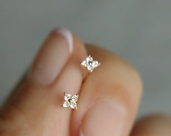 May include: A pair of gold earrings with a small flower design set with clear diamonds. The earrings are held in a hand.