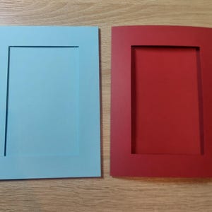 Puede incluir: Dos marcos de fotos rectangulares, uno azul claro y otro rojo, se muestran uno al lado del otro. Cada marco tiene un rectángulo interior empotrado. Los marcos están sobre una superficie de madera marrón claro.