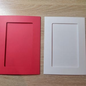 Puede incluir: Dos marcos de fotos rectangulares, uno rojo brillante y otro blanco, se muestran uno al lado del otro. Cada marco tiene una abertura rectangular empotrada en el centro, diseñada para contener una fotografía o una obra de arte. Los marcos están sobre una superficie de madera clara.