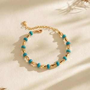 Könnte beinhalten: Ein goldfarbenes Armband mit abwechselnd türkisfarbenen und goldfarbenen zylindrischen Perlen. Das Armband liegt auf einer weißen Oberfläche, mit Schatten und getrockneten Pflanzen im Hintergrund. Es hat einen Karabinerverschluss.