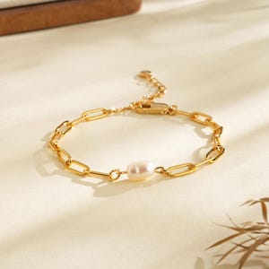 Peut inclure: Bracelet en chaîne dorée avec une perle ovale au centre. Le bracelet présente un design de chaîne trombone et une fermeture à fermoir mousqueton. Le bijou est présenté sur une surface crème avec des ombres douces.