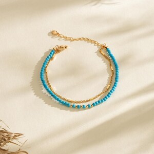 Puede incluir: Pulsera de dos hilos con pequeñas cuentas de color turquesa y eslabones de cadena dorados. La pulsera tiene un diseño delicado con un pequeño dije redondo en el extremo. La joya se exhibe sobre una superficie color crema.