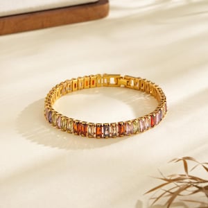 May include: A gold bracelet featuring a series of rectangular, multi-coloured gemstones. The gemstones are in shades of red, orange, yellow, green, purple, and pink. The bracelet is on a white surface with shadows.