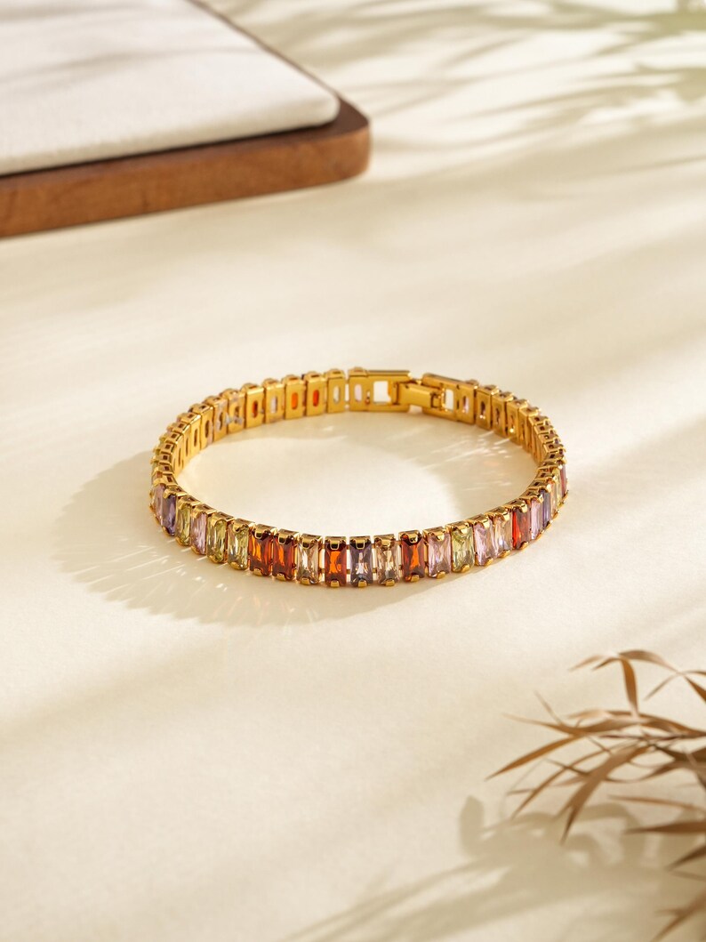 May include: A gold bracelet featuring a series of rectangular, multi-coloured gemstones. The gemstones are in shades of red, orange, yellow, green, purple, and pink. The bracelet is on a white surface with shadows.