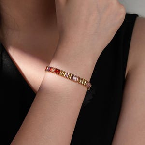 May include: A gold bracelet with a line of colourful, rectangular gemstones. The gemstones are in shades of red, orange, yellow, and pink. The bracelet is worn on a wrist against a black background.