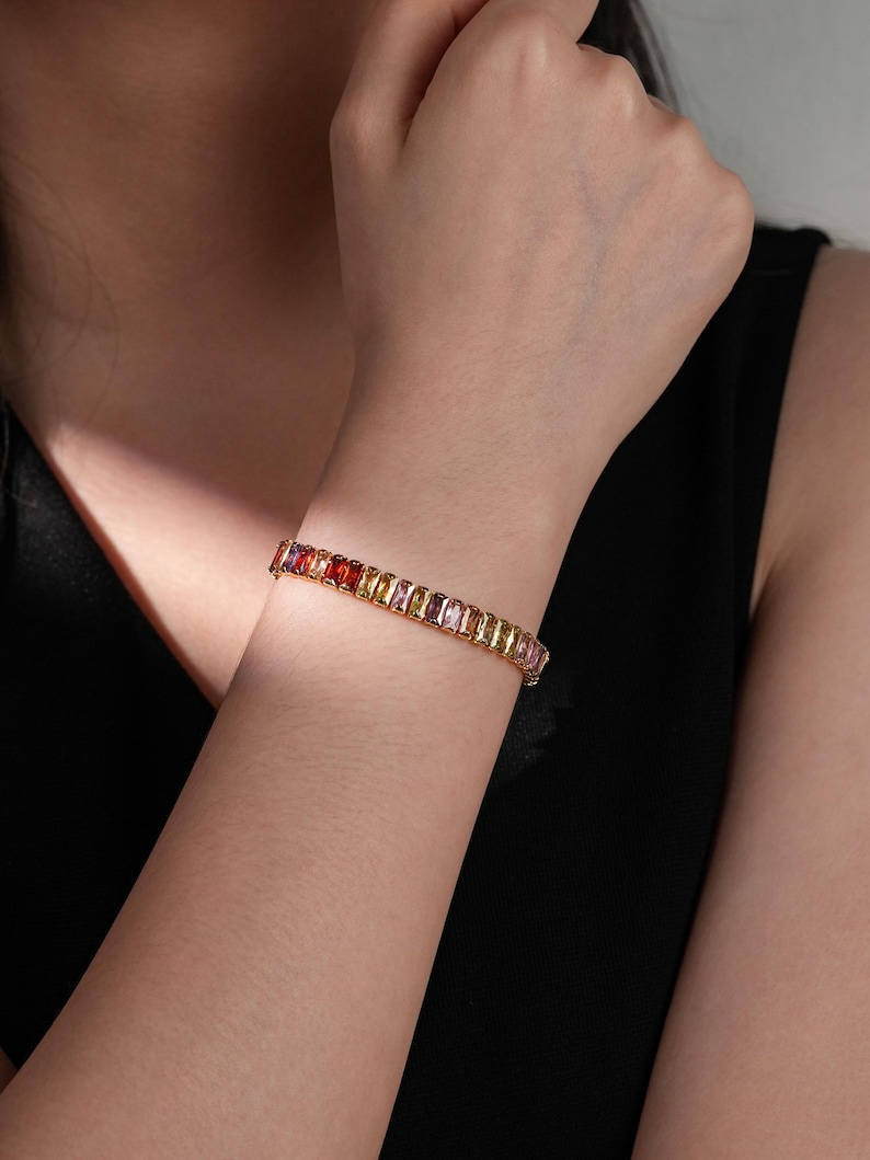 May include: A gold bracelet with a line of colourful, rectangular gemstones. The gemstones are in shades of red, orange, yellow, and pink. The bracelet is worn on a wrist against a black background.