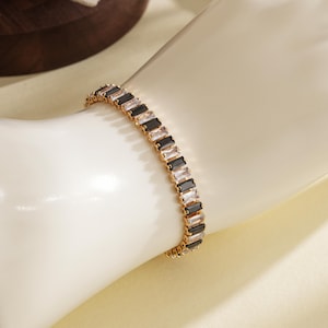 May include: A gold bracelet featuring alternating black and clear rectangular gemstones. The bracelet is presented on a mannequin arm, highlighting its design. The gemstones are arranged in a linear pattern, offering a contemporary and refined aesthetic.