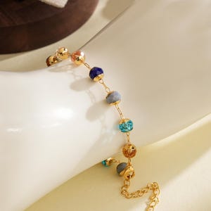May include: Gold bracelet with a chain and a series of colourful, spherical beads. The beads are in shades of blue, teal, orange, and grey. The bracelet is displayed on a white surface, highlighting its delicate design and vibrant colours.