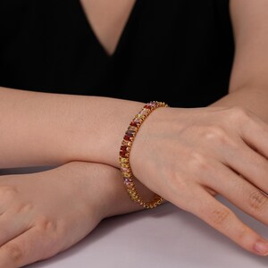 May include: A gold bracelet featuring a line of multi-coloured rectangular gemstones. The gemstones are in shades of red, orange, yellow, pink, and purple. The bracelet is worn on a wrist against a black top.