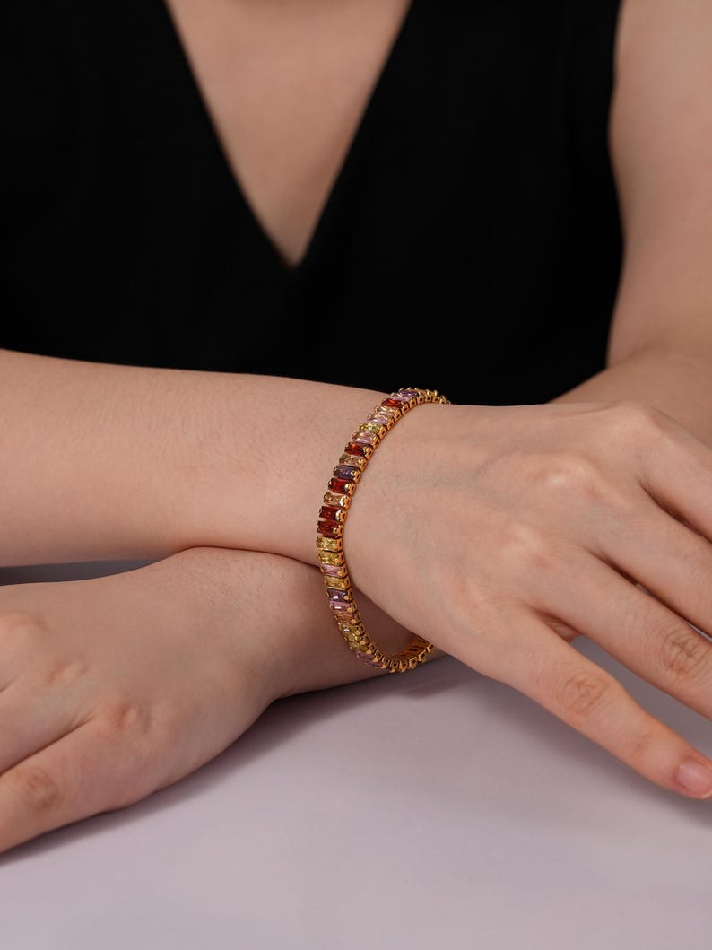 May include: A gold bracelet featuring a line of multi-coloured rectangular gemstones. The gemstones are in shades of red, orange, yellow, pink, and purple. The bracelet is worn on a wrist against a black top.