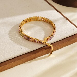 May include: Gold-coloured bracelet with a row of colourful rectangular gemstones. The bracelet has a secure clasp and is displayed on a white surface with a wooden border. The gemstones include shades of orange, yellow, pink, and purple.