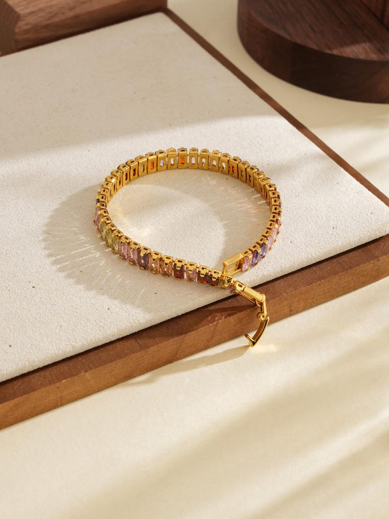 May include: Gold-coloured bracelet with a row of colourful rectangular gemstones. The bracelet has a secure clasp and is displayed on a white surface with a wooden border. The gemstones include shades of orange, yellow, pink, and purple.