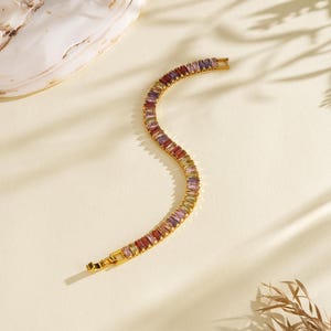 May include: A gold bracelet featuring a line of multi-coloured rectangular gemstones. The gemstones are in shades of red, purple, yellow, and green. The bracelet is on a cream surface with shadows and a seashell.